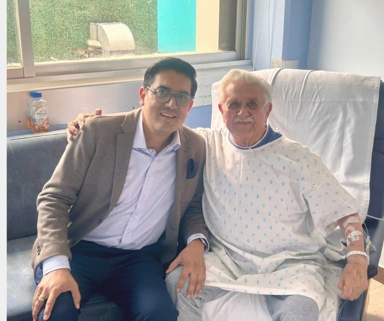 A man in a beige blazer and glasses sits beside an elderly patient in a hospital gown on a medical bed